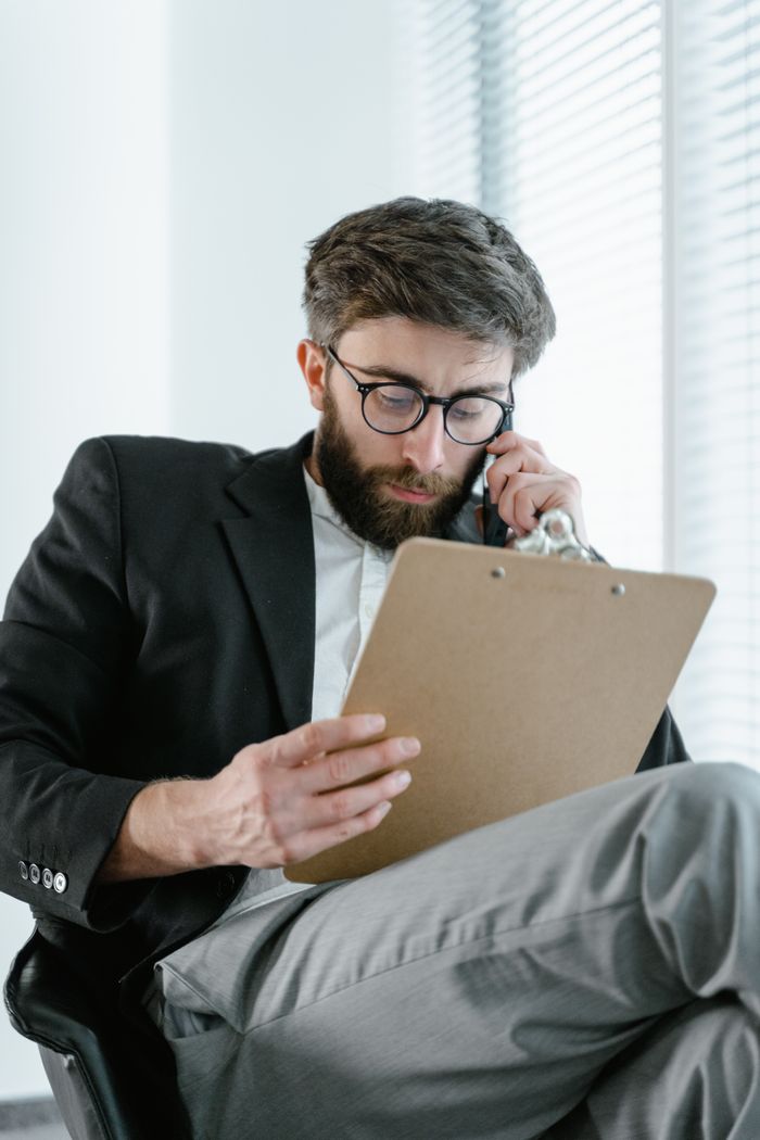 A bearded businessman in an office setting examines paperwork while on a phone call.